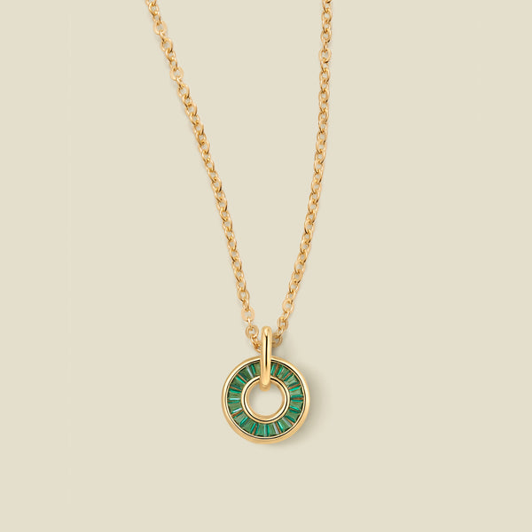 A gold chain necklace with a circular pendant featuring dark green baguette-cut stones around a hollow center, displayed against a solid light beige background.