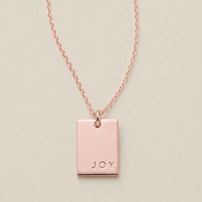 A rose gold rectangular pendant necklace engraved with the word 'JOY' in the bottom right corner, displayed on a light beige background.