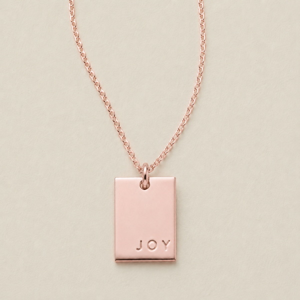 A rose gold rectangular pendant necklace engraved with the word 'JOY' in the bottom right corner, displayed on a light beige background.