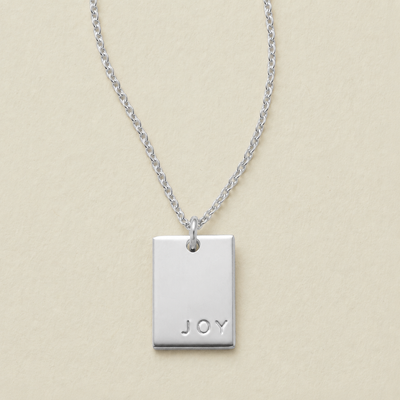A silver rectangular pendant necklace engraved with the word 'JOY' in the bottom right corner, on a light beige textured background.