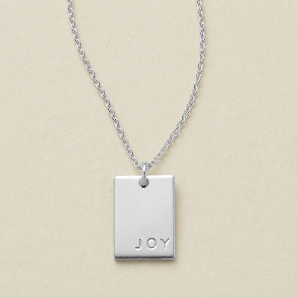 A silver rectangular pendant necklace engraved with the word 'JOY' in the bottom right corner, on a light beige textured background.