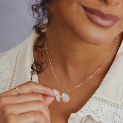 A close-up lifestyle shot of a model wearing a delicate gold chain necklace with an open oval locket, being held by her finger.