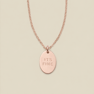 A rose gold oval pendant necklace engraved with the words 'ITS FINE' on a matching chain, displayed against a solid light beige background.