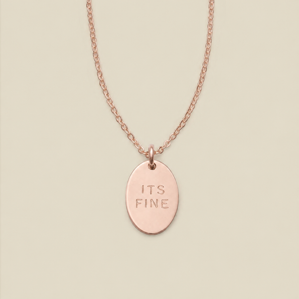 A rose gold oval pendant necklace engraved with the words 'ITS FINE' on a matching chain, displayed against a solid light beige background.