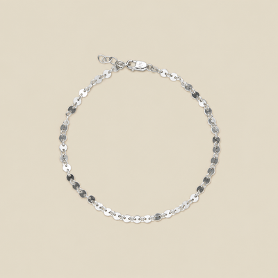 A delicate silver chain bracelet with flat, polished circular links, displayed in a circular shape on a solid beige background.