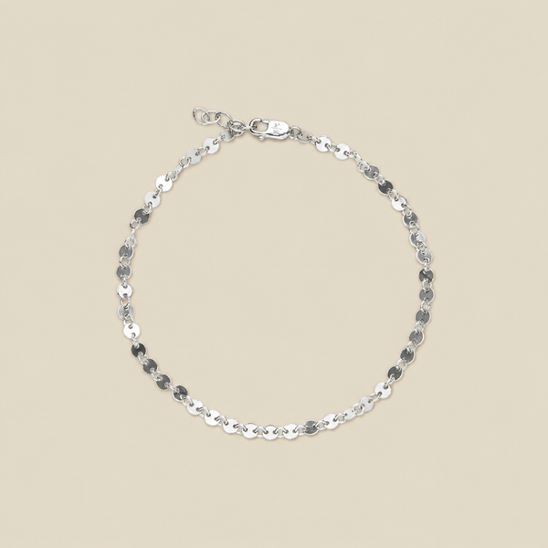 A delicate silver chain bracelet with flat, polished circular links, displayed in a circular shape on a solid beige background.