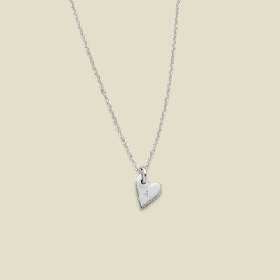 A delicate silver chain necklace with a small, polished heart-shaped pendant engraved with an uppercase letter 'A', displayed against a solid light beige background.