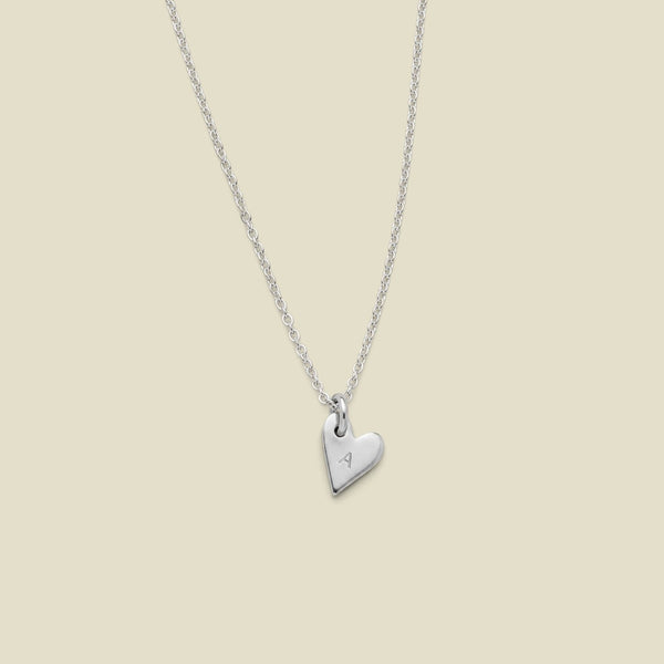 A delicate silver chain necklace with a small, polished heart-shaped pendant engraved with an uppercase letter 'A', displayed against a solid light beige background.