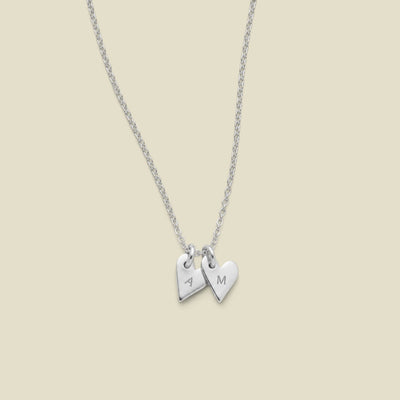 A dainty silver chain necklace with two small heart charms, one engraved with the letter 'A' and the other with 'M', displayed against a solid light beige background.