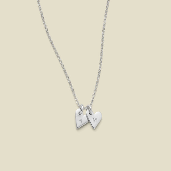 A dainty silver chain necklace with two small heart charms, one engraved with the letter 'A' and the other with 'M', displayed against a solid light beige background.
