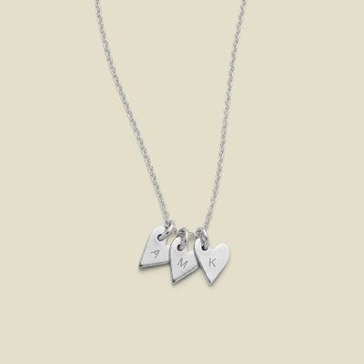 A dainty silver necklace with three small  heart charms, each engraved with a different letter (A, M, K), displayed on a light beige background.