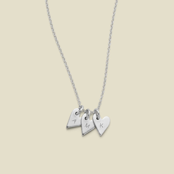 A dainty silver necklace with three small  heart charms, each engraved with a different letter (A, M, K), displayed on a light beige background.