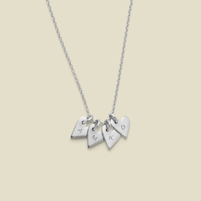 A silver necklace with four small heart-shaped charms engraved with the letters A, M, K, and D, displayed against a solid light beige background.