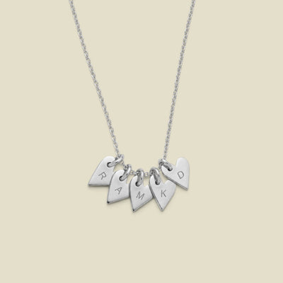 A silver necklace with five small heart-shaped charms engraved with the letters R, A, M, K, and D, displayed against a solid light beige background.
