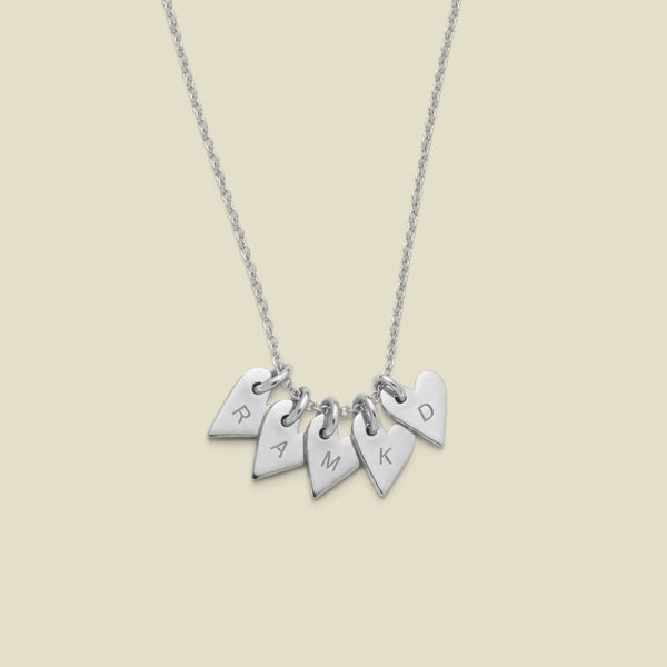 A silver necklace with five small heart-shaped charms engraved with the letters R, A, M, K, and D, displayed against a solid light beige background.