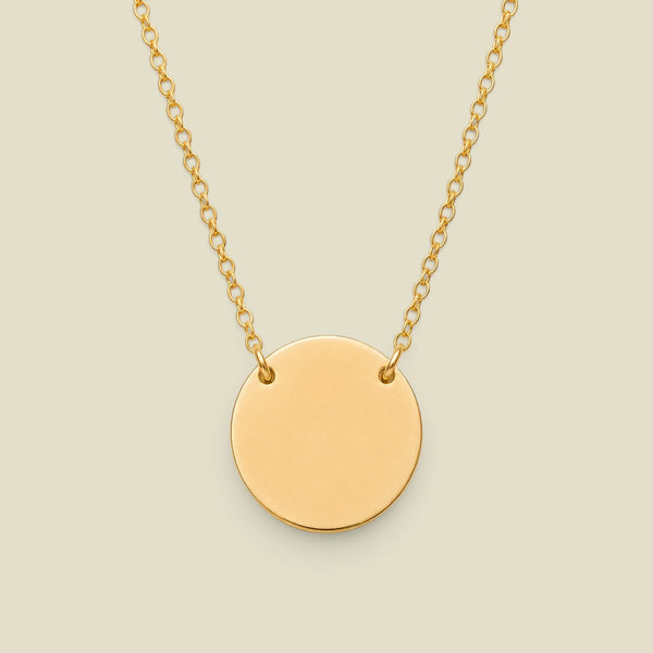 A minimalist gold disc pendant necklace attached to a delicate gold chain at two spots on the upper half of the disc, centered against a solid light beige background.