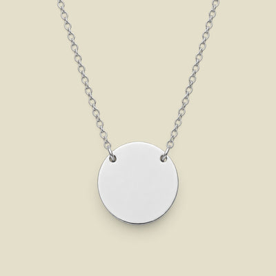 A silver disc pendant necklace connected to a delicate silver chain in two spots, presented against a solid light beige background.