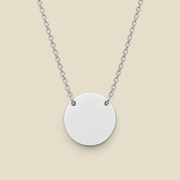 A silver disc pendant necklace connected to a delicate silver chain in two spots, presented against a solid light beige background.