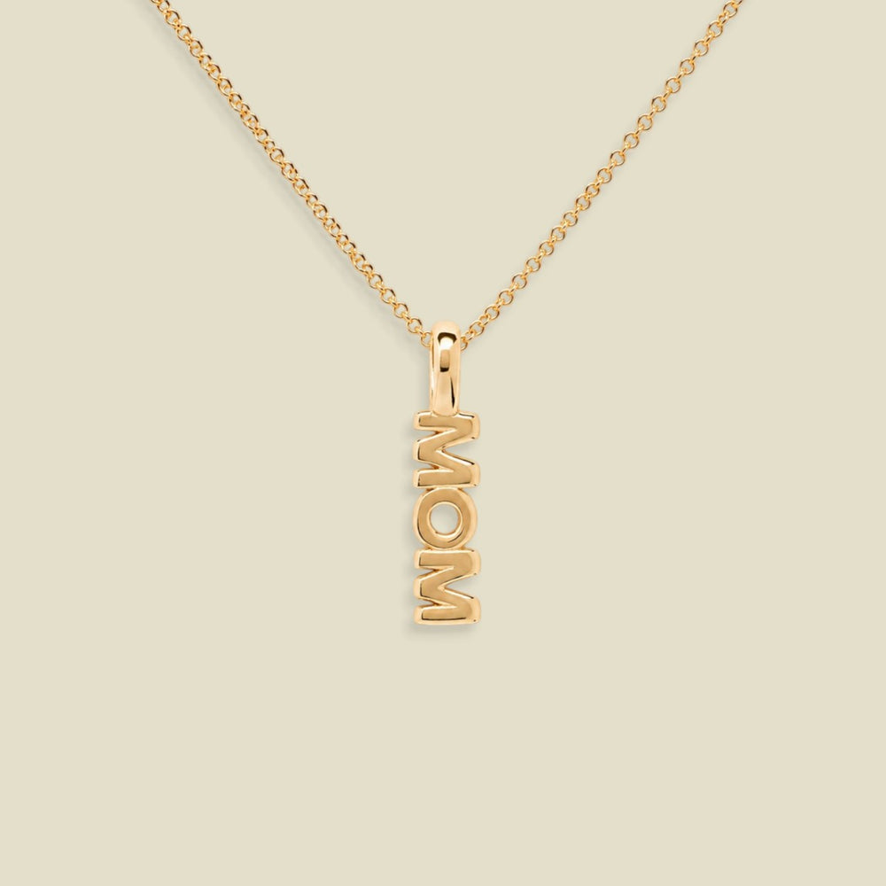 A gold chain necklace with a vertical pendant spelling 'MOM', shown on a plain light beige background.
