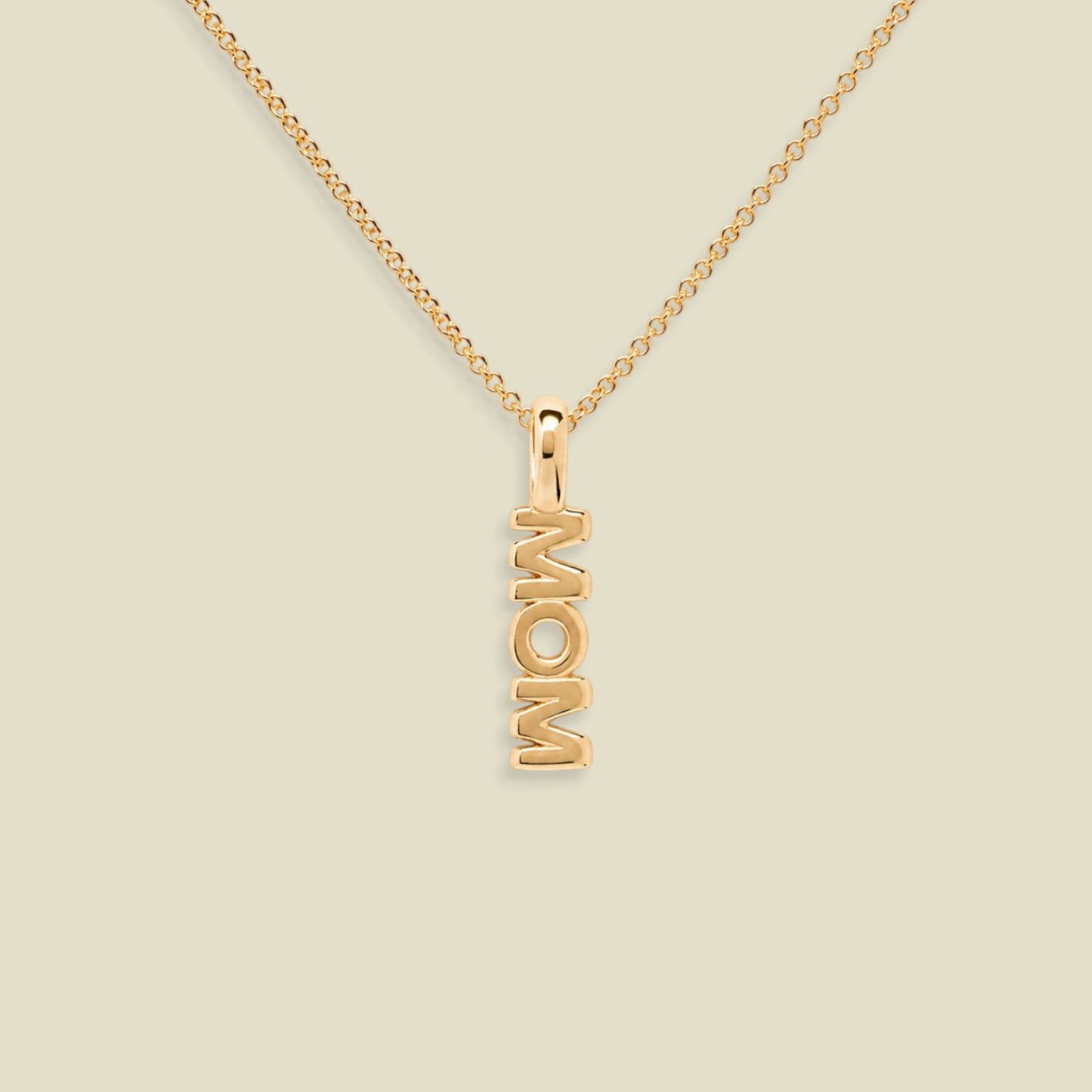 A gold chain necklace with a vertical pendant spelling 'MOM', shown on a plain light beige background.