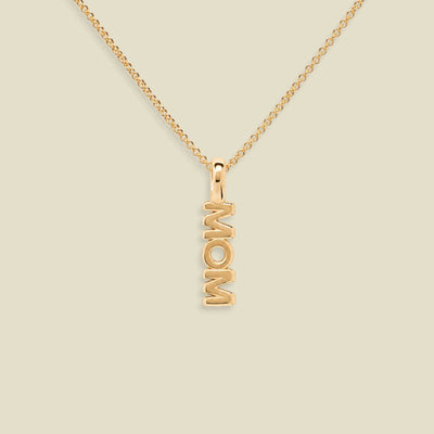 A gold chain necklace with a vertical pendant spelling 'MOM', shown on a plain light beige background.
