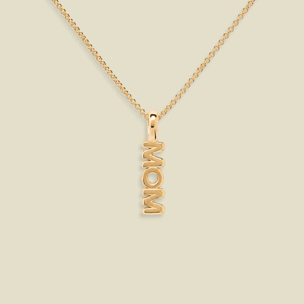 A gold chain necklace with a vertical pendant spelling 'MOM', shown on a plain light beige background.