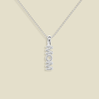 A silver necklace with a vertical pendant spelling 'MOM', displayed on a clean beige background.