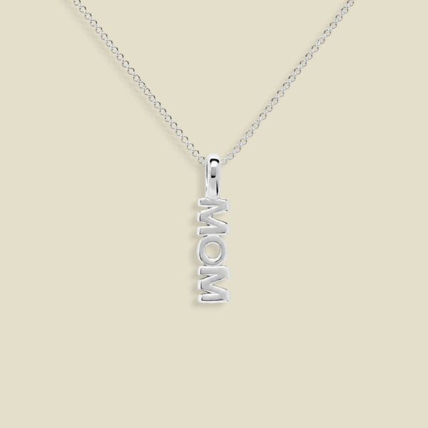 A silver necklace with a vertical pendant spelling 'MOM', displayed on a clean beige background.