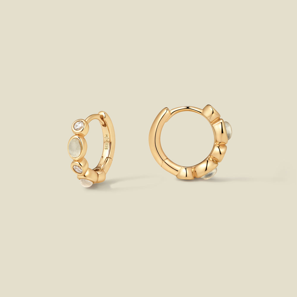 Pair of gold hoop earrings with white and clear bezel set stones against a neutral background.