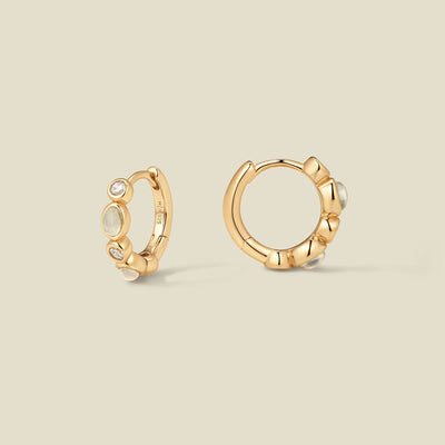 Pair of gold hoop earrings with white and clear bezel set stones against a neutral background.