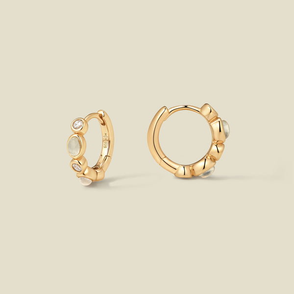 Pair of gold hoop earrings with white and clear bezel set stones against a neutral background.