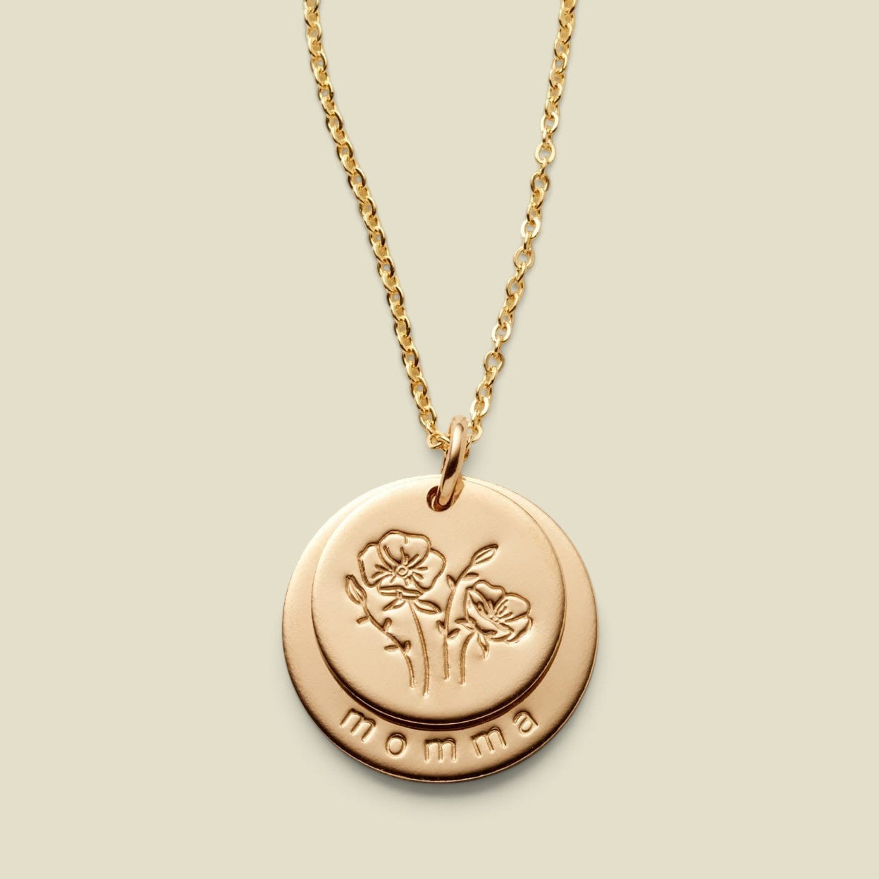 A gold disc pendant necklace featuring two overlapping discs on one jump ring, one with an engraved floral design, and the other larger disc underneath with the word 'momma' along the  bottom, displayed against a solid light beige background.