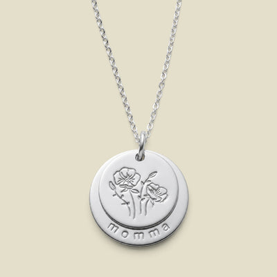 A silver round disc pendant necklace with two overlapping discs on one jump ring, with the first being engraved with a flower design, and the larger disc underneath stamped with the word 'momma' along the bottom edge, displayed on a light beige background.