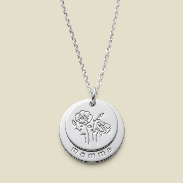 A silver round disc pendant necklace with two overlapping discs on one jump ring, with the first being engraved with a flower design, and the larger disc underneath stamped with the word 'momma' along the bottom edge, displayed on a light beige background.