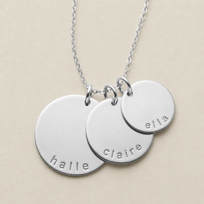 A silver chain necklace with three overlapping round disc pendants engraved with the names 'halle', 'claire', and 'ella' on a light beige background.