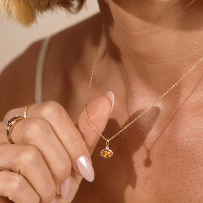 A close-up lifestyle shot of a woman wearing a delicate gold chain necklace with a small gold oval-shaped pendant set with a faceted orange gemstone. The image highlights the jewelry on her neck and chest, with her hand gently touching the chain.