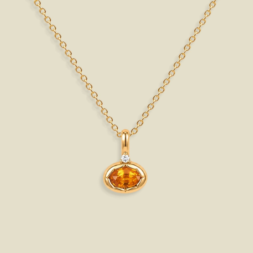 A gold chain necklace with a small oval orange gemstone pendant in a gold bezel setting, accented by a tiny clear stone on the top, shown against a solid light beige background.