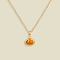 A gold chain necklace with a small oval orange gemstone pendant in a gold bezel setting, accented by a tiny clear stone on the top, shown against a solid light beige background.