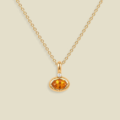 A gold chain necklace with a small oval orange gemstone pendant in a gold bezel setting, accented by a tiny clear stone on the top, shown against a solid light beige background.