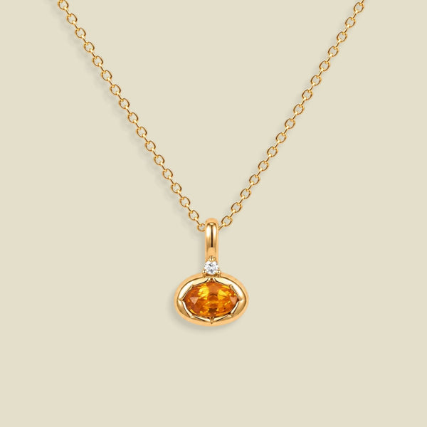 A gold chain necklace with a small oval orange gemstone pendant in a gold bezel setting, accented by a tiny clear stone on the top, shown against a solid light beige background.