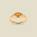 A gold signet ring with an amber center oval bezel-set gemstone, and two small clear accent stones on either side, set against a solid beige background.