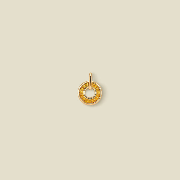 A gold circular pendant featuring golden yellow baguette-cut stones around a hollow center, displayed against a solid light beige background.