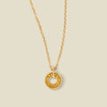 A gold chain necklace with a circular pendant featuring amber baguette-cut stones around a hollow center, displayed against a solid light beige background.