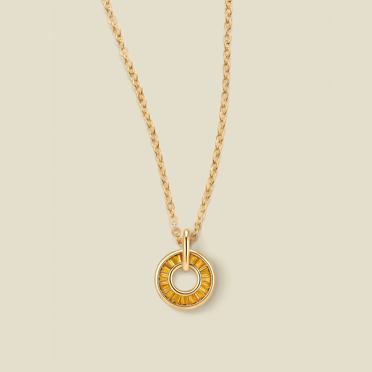 A gold chain necklace with a circular pendant featuring amber baguette-cut stones around a hollow center, displayed against a solid light beige background.
