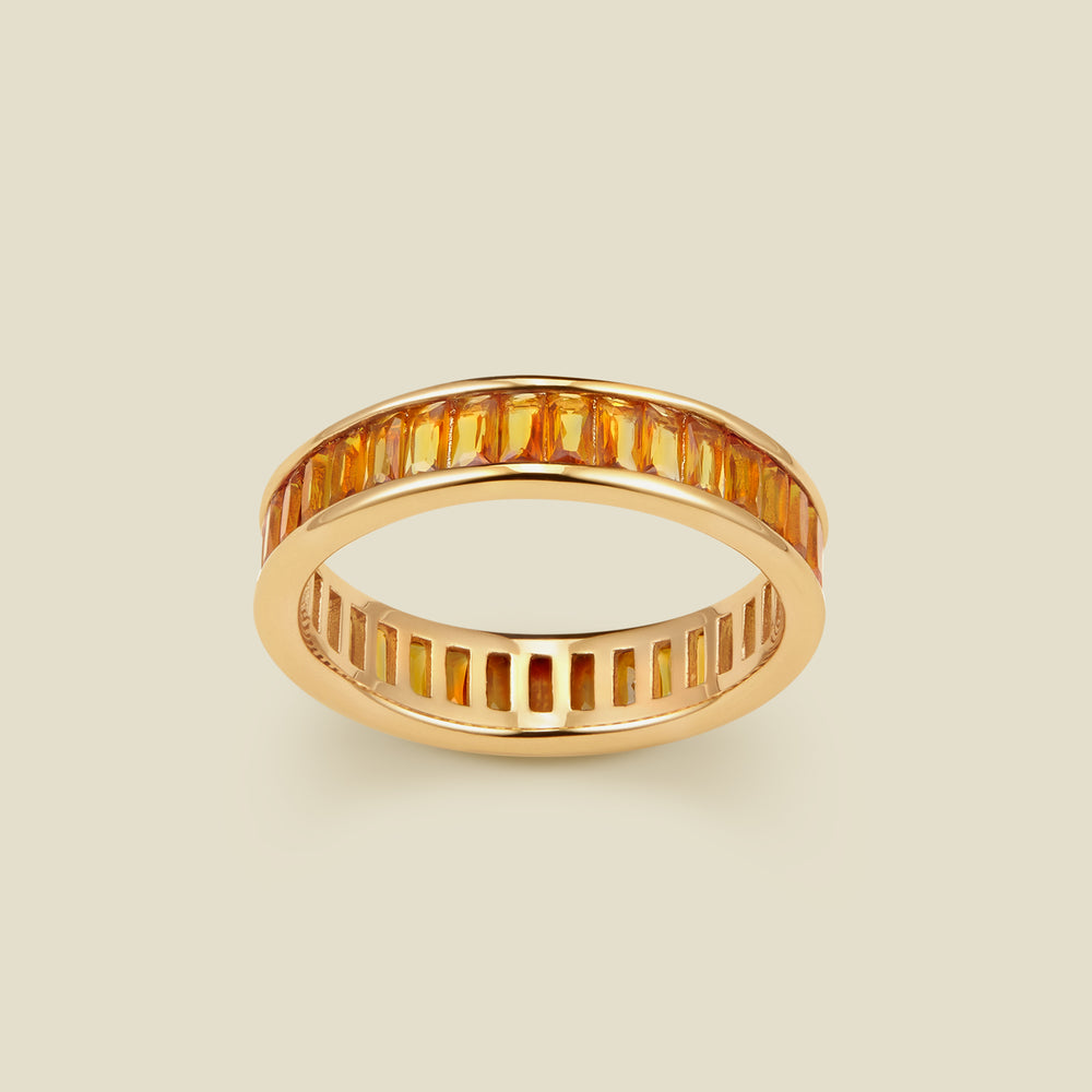 A gold eternity ring featuring a continuous channel setting of baguette-cut amber gemstones, shown on a solid light beige background.