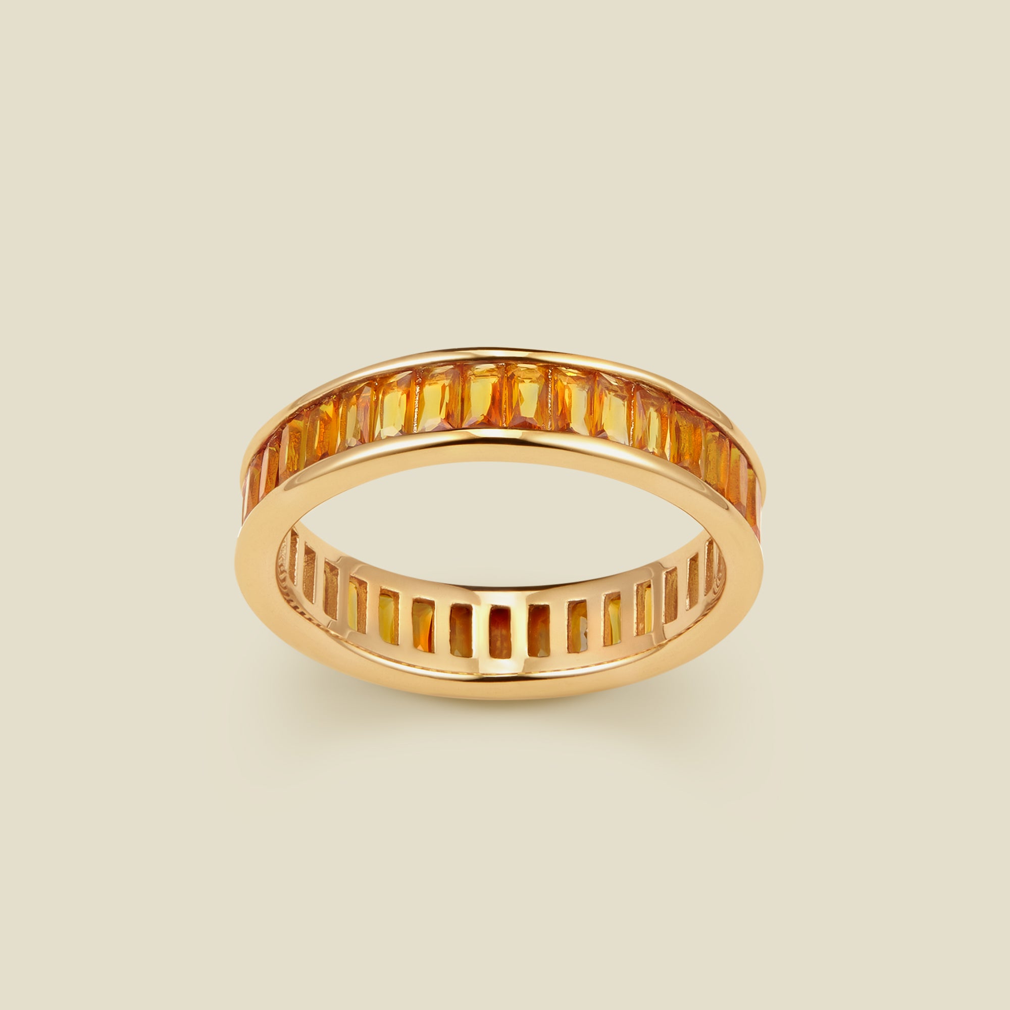 A gold eternity ring featuring a continuous channel setting of baguette-cut amber gemstones, shown on a solid light beige background.