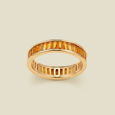 A gold eternity ring featuring a continuous channel setting of baguette-cut amber gemstones, shown on a solid light beige background.