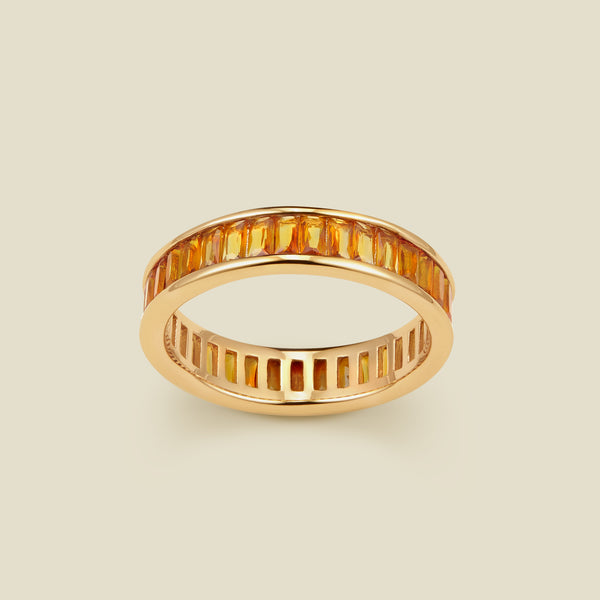 A gold eternity ring featuring a continuous channel setting of baguette-cut amber gemstones, shown on a solid light beige background.