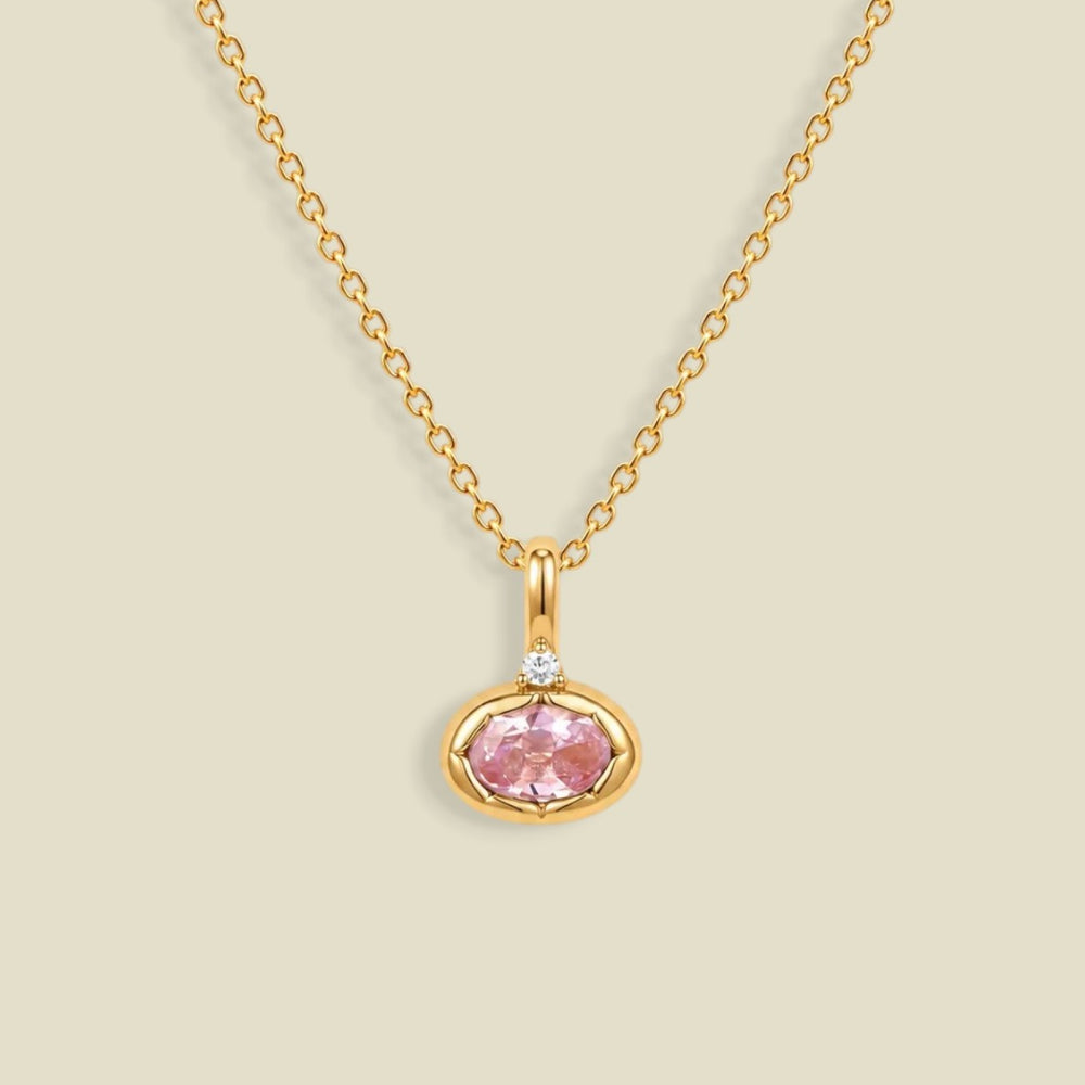 A delicate gold chain necklace with a small oval pendant featuring a pink gemstone and a tiny clear accent stone above it, set against a solid light beige background.