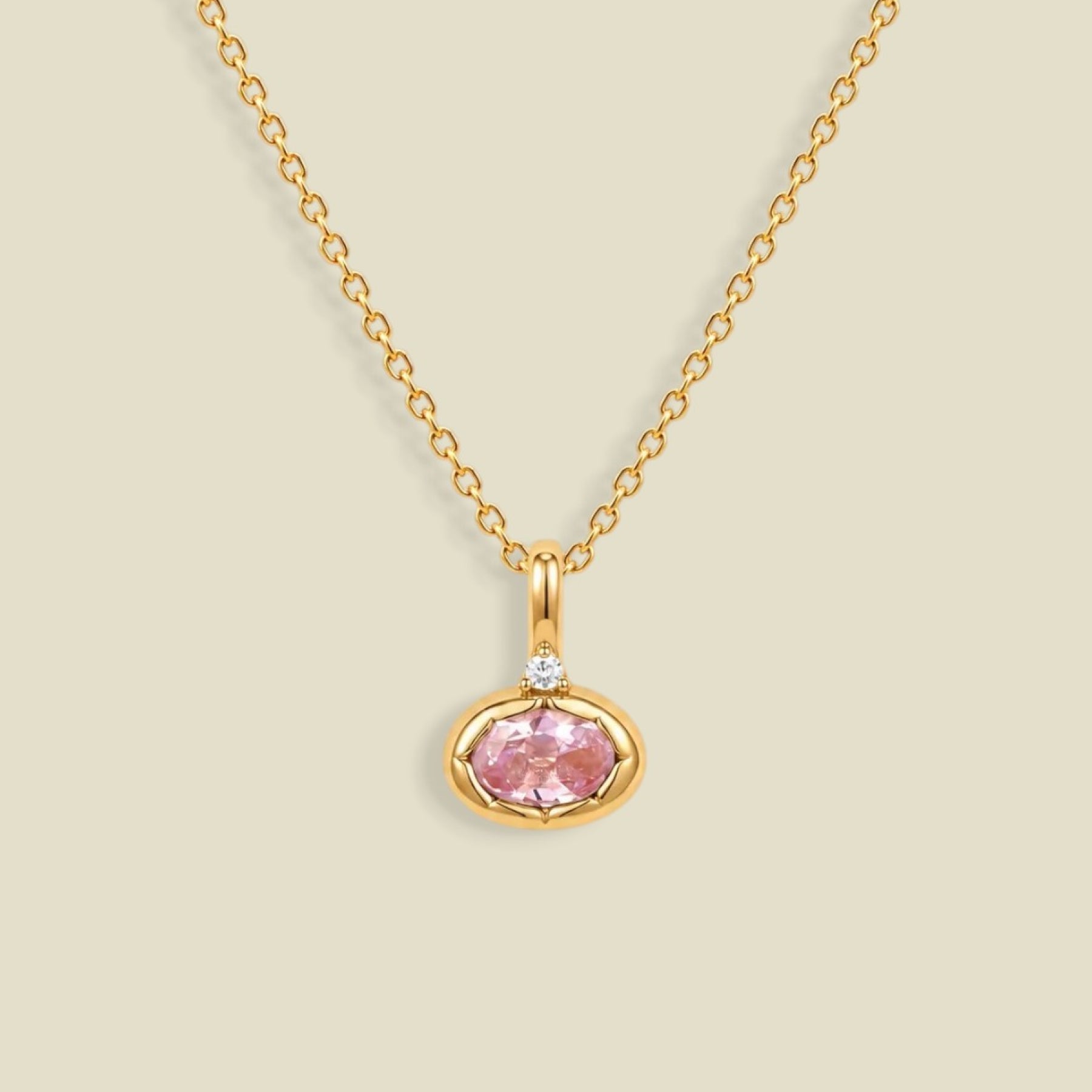 A delicate gold chain necklace with a small oval pendant featuring a pink gemstone and a tiny clear accent stone above it, set against a solid light beige background.